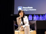 A woman sits on a stage at an awards program, holding a microphone and speaking to an audience